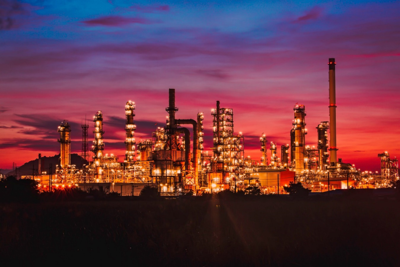 A large oil refinery lit up against an evening sky in shades of pink and purple shows the sheer scale of the plant, highlighting the importance of strategic refinery acquisitions for success.