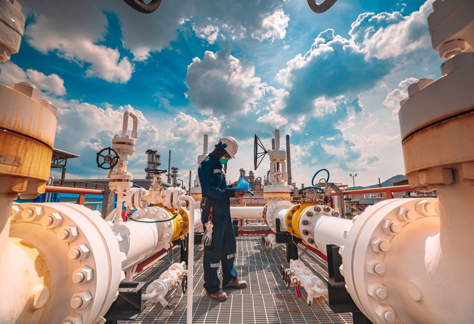 A man standing between large white and yellow pipes with a bright blue sky in the background, highlighting the complexity of the refinery acquisition process.