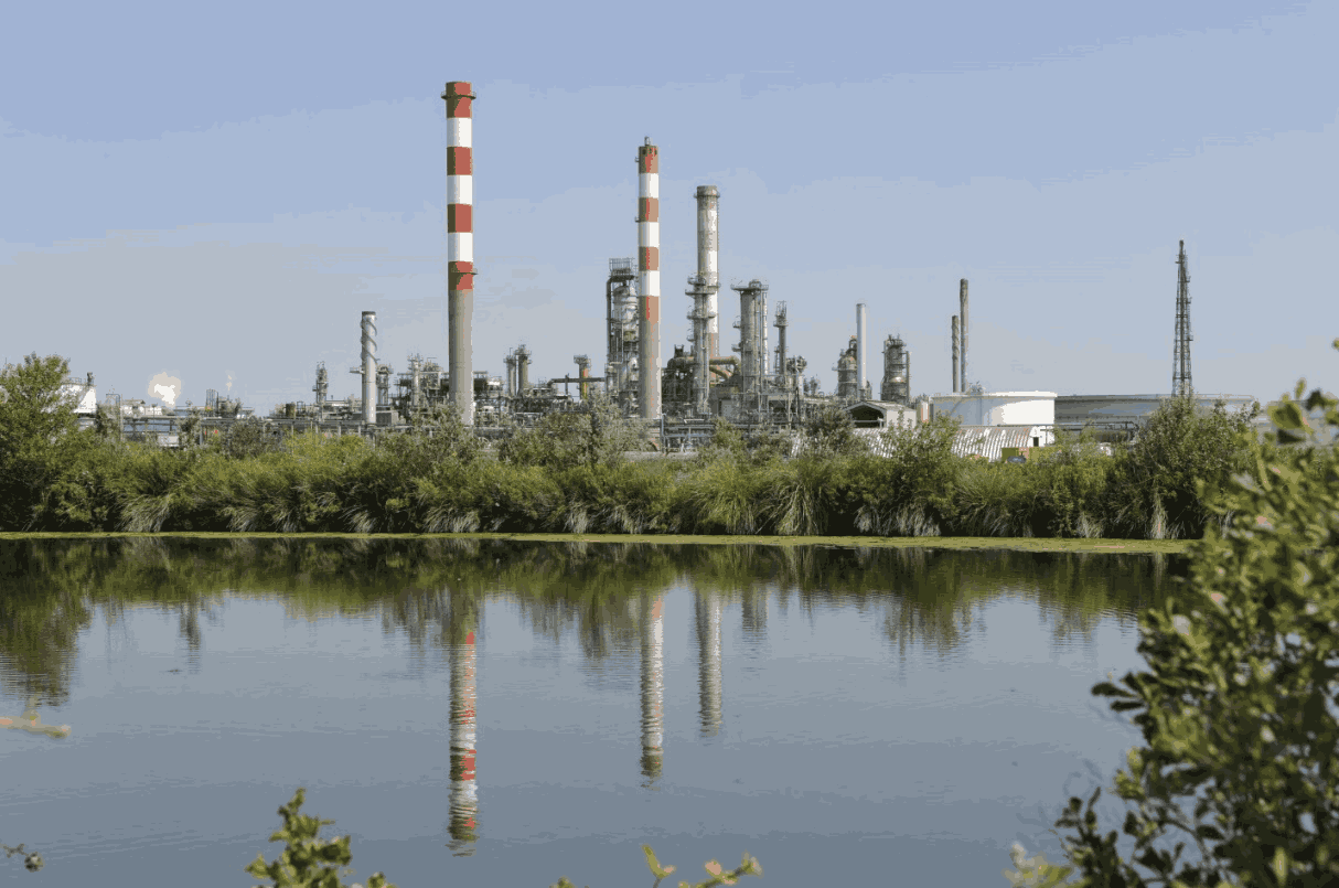 An oil refinery reflected in a body of water surrounded by foliage, symbolizing the need for efficiency in refinery operations.