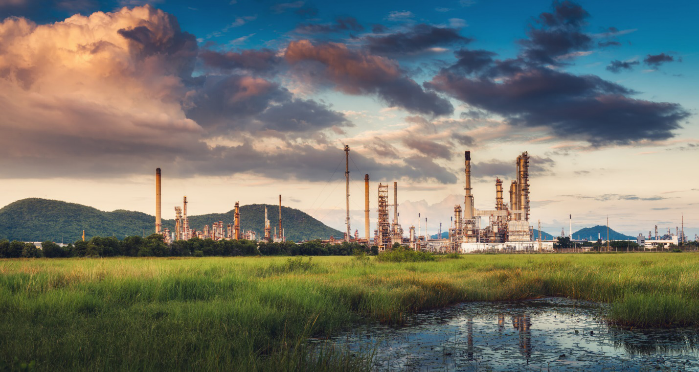 Grass grows in this extensive area leading up to a large oil refinery setup under a cloudy evening sky, showing the sheer scale that requires efficiency in refinery operations.