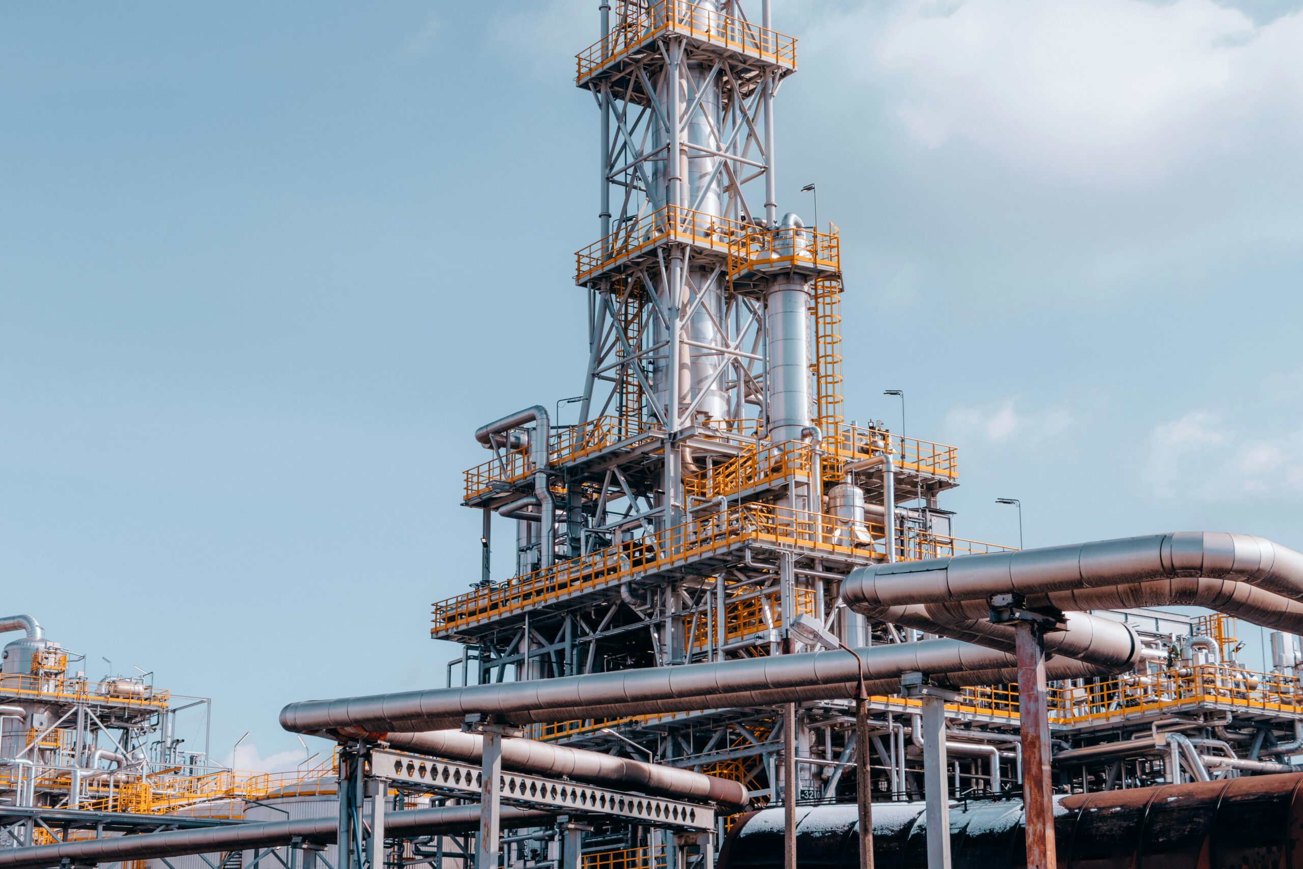 A close-up of part of an oil refinery against a light blue sky with scattered clouds