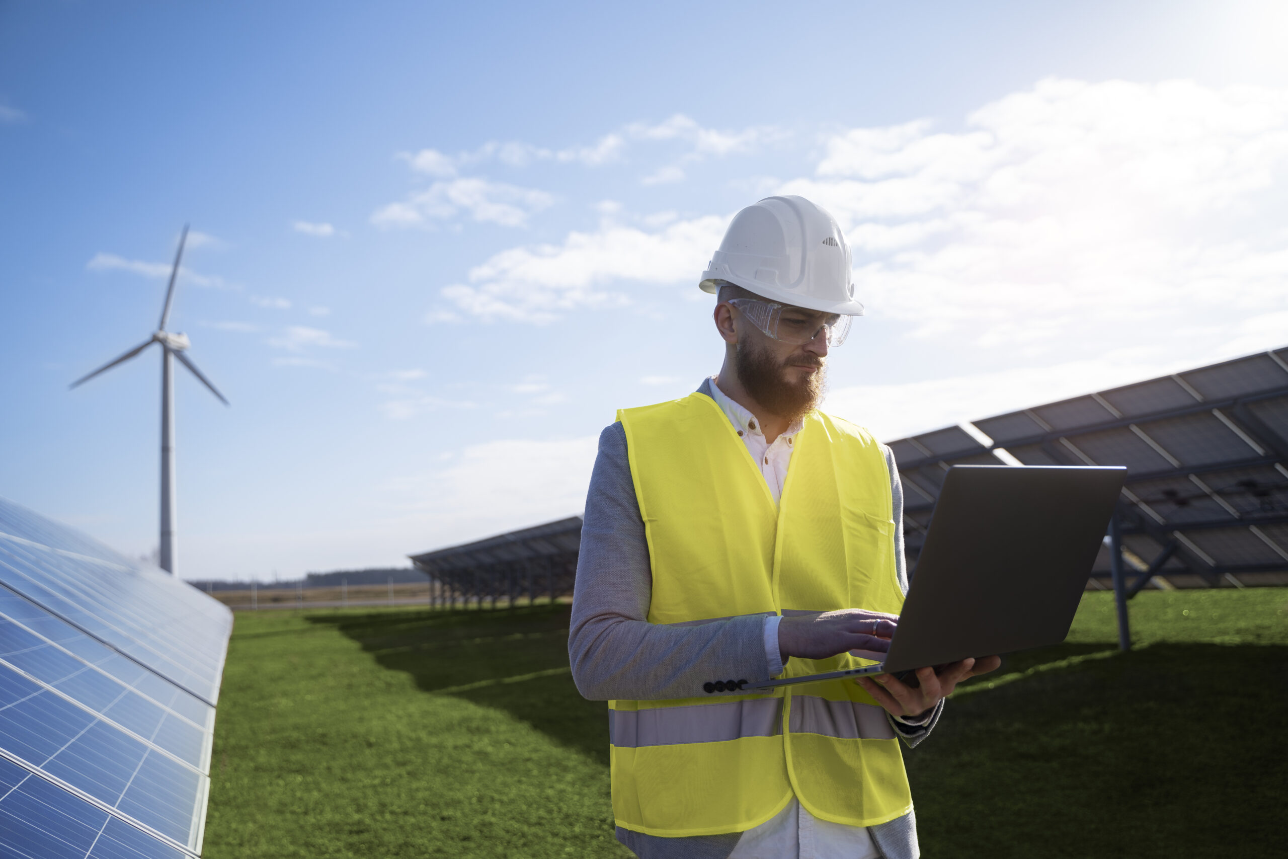 A man wearing a hard hat looking at a laptop stands next to a solar panel with a wind turbine in the background